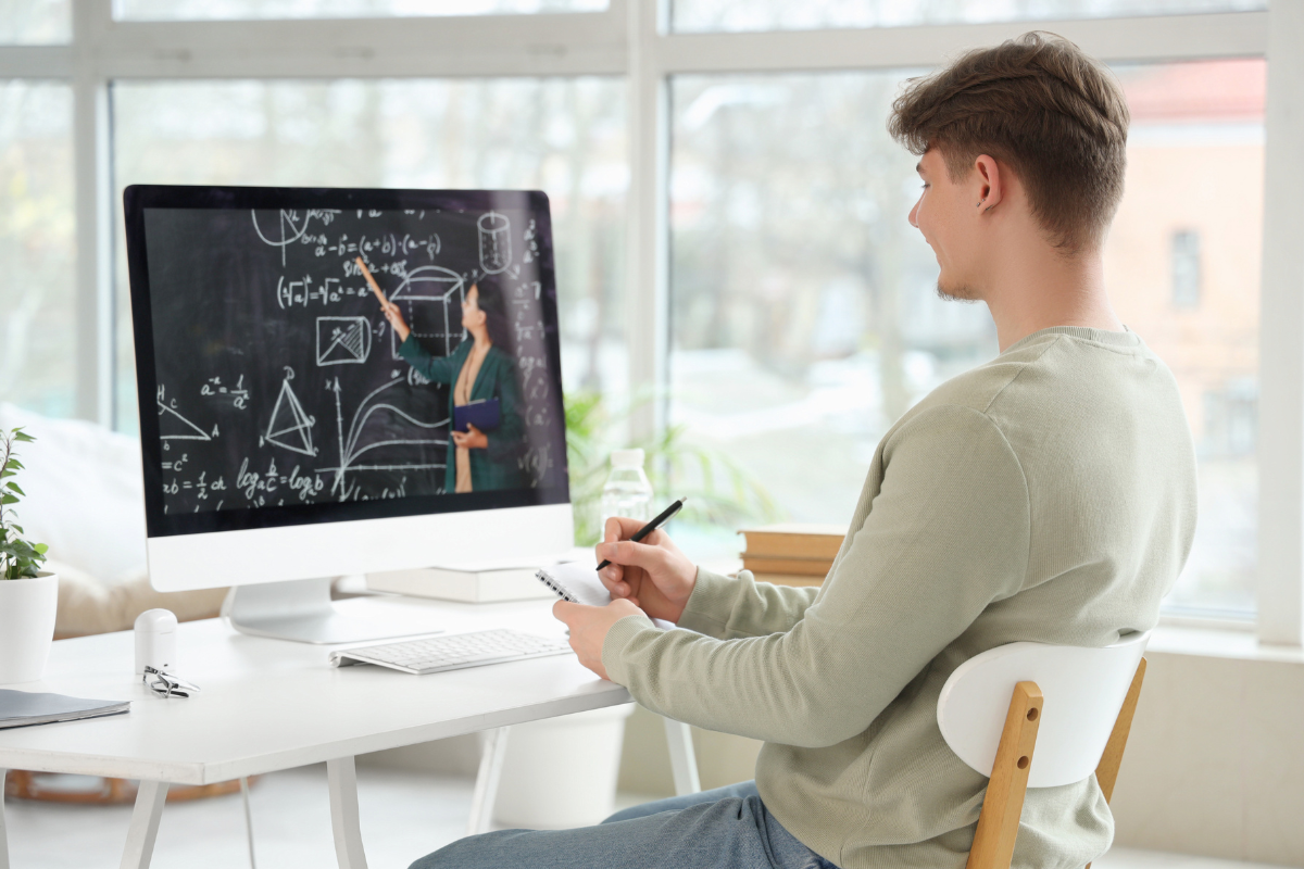 A college student takes notes while using AI courseware for calculus on a computer screen. The screen displays a teacher pointing to mathematical formulas and diagrams on a chalkboard.