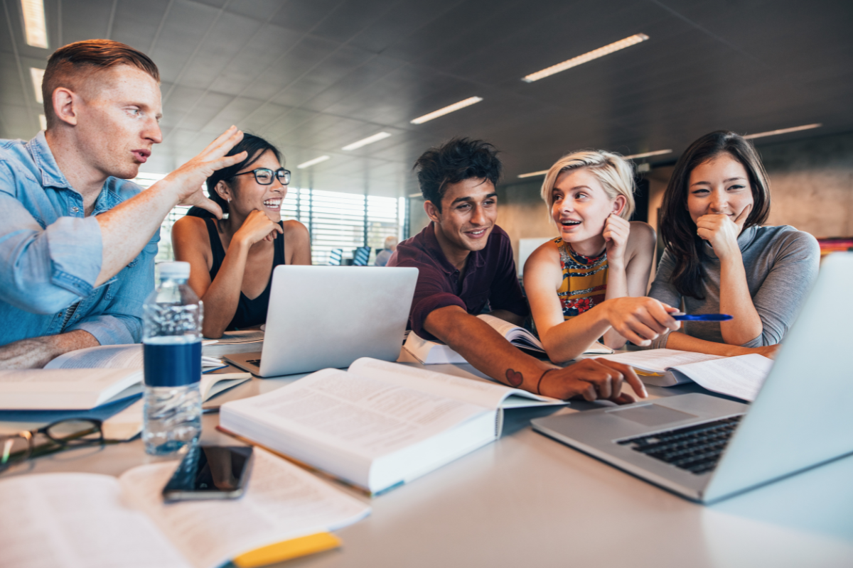 A student in a blue shirt speaks about AI for student success while gesturing to a group of students seated around a table with laptops, notebooks, and water bottles in a classroom.