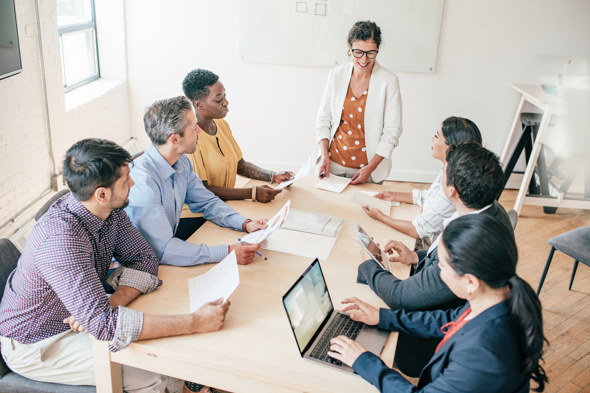 A female faculty member stands at a wooden table, leading a meeting on AI leadership in higher education with faculty seated around laptops and papers in a bright office classroom.