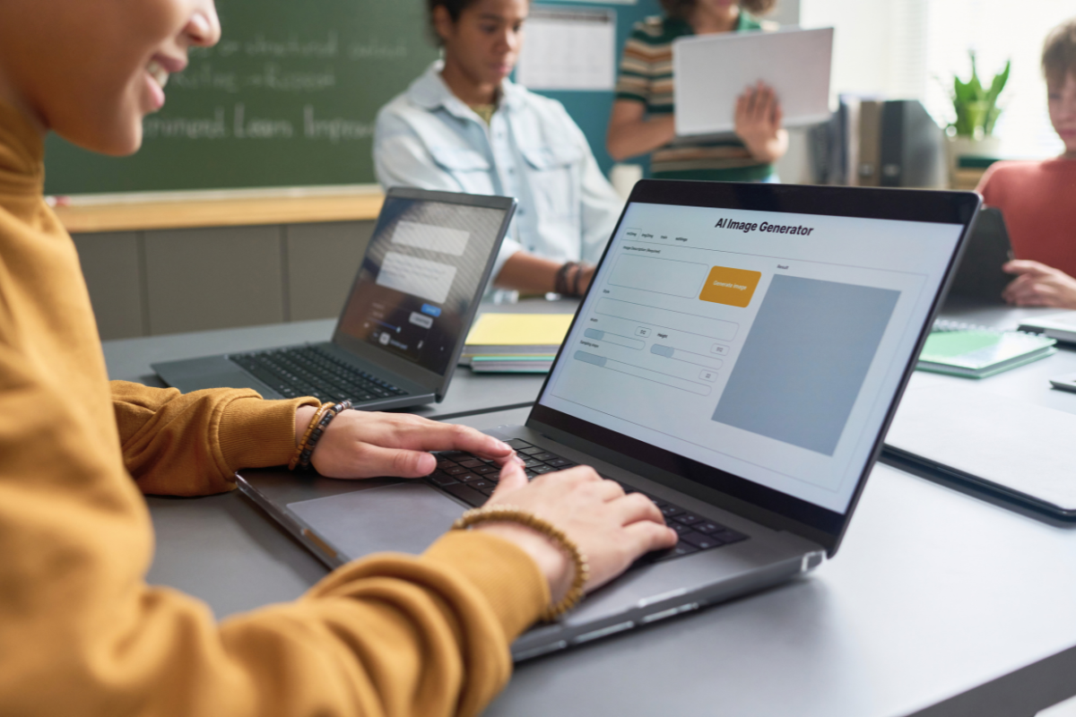 A student typing on an open laptop at a classroom table, demonstrating authentic learning with AI, with another student standing in the background and a laptop screen showing an AI interface.