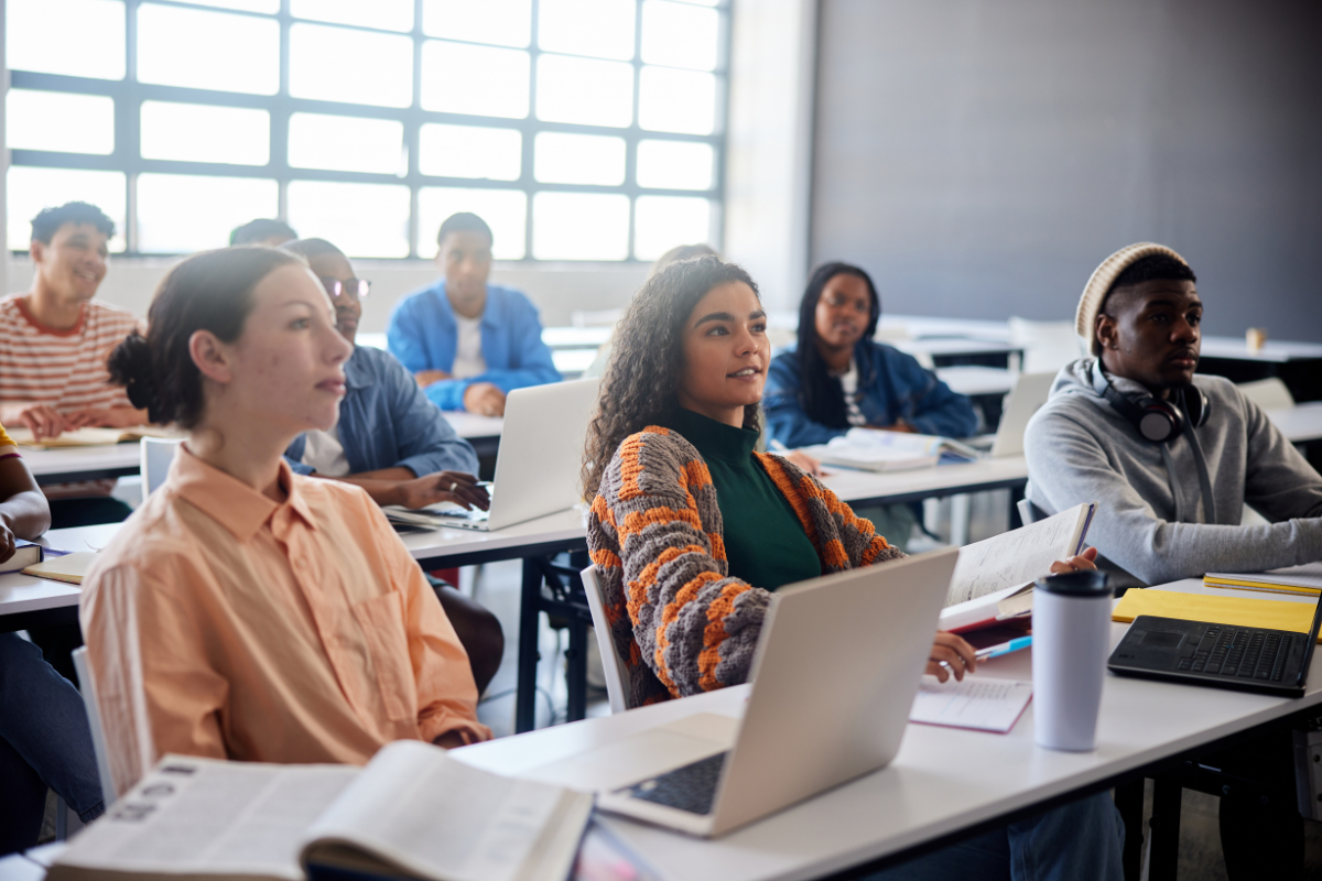 Students sit at desks in a digital classroom, with laptops and books open in front of them. Large windows let in natural light from above.