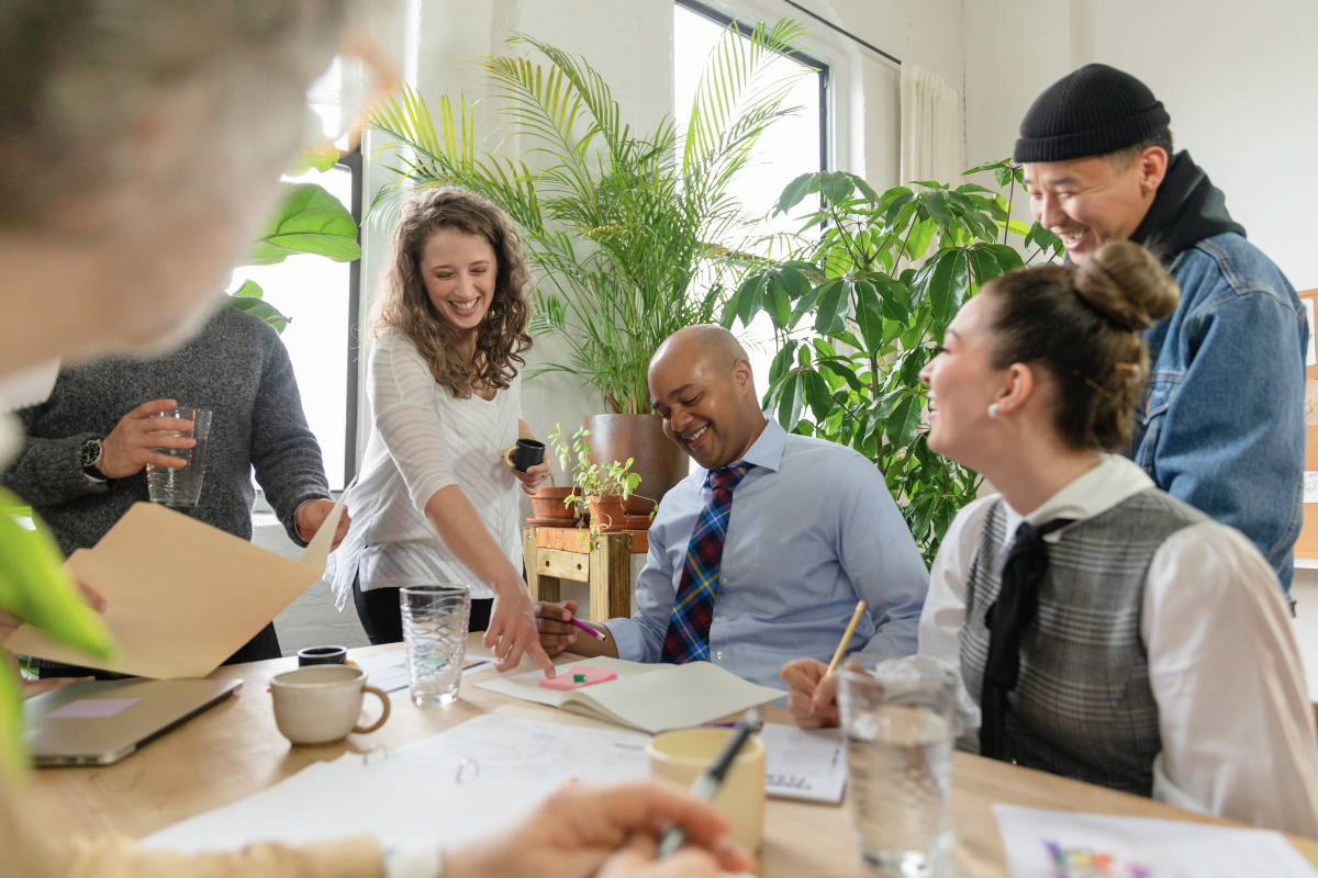 A group of faculty collaborating at a table during a meeting on gateway course optimization, smiling and writing with cups and papers, while a man in a beanie stands nearby in a bright room with green plants.
