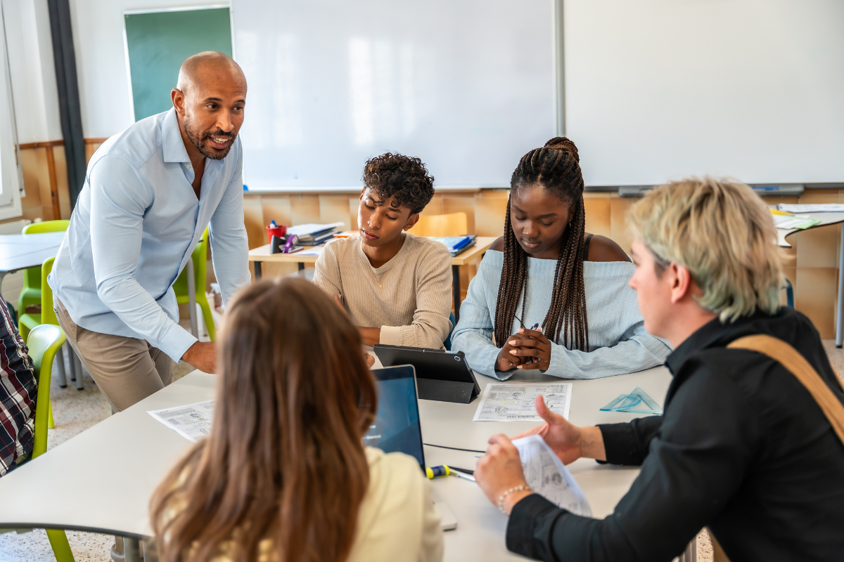A teacher stands while listening to learners with a small group of students seated at desks in a classroom, with hands-on discussion during a lesson.
