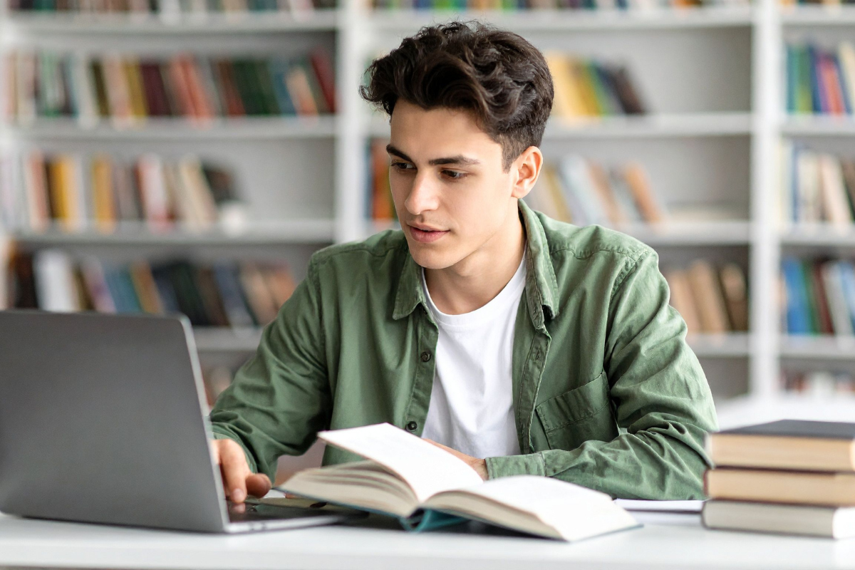 A college student wearing a green shirt is sitting at a desk with a laptop and an open book in front of them practicing metacognition in college teaching. Bookshelves filled with books are visible in the background.