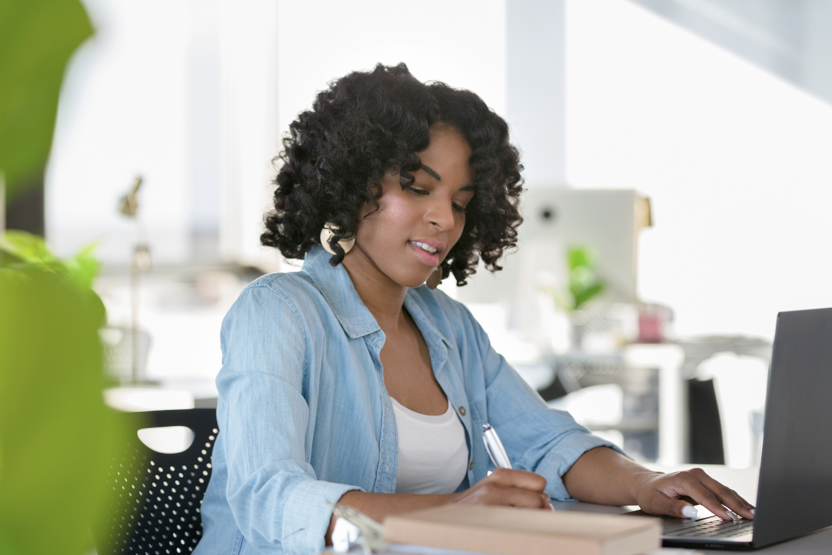A student with curly hair works at a desk, typing on a laptop and writing on papers in a bright office with soft light and blurred greenery in the foreground.