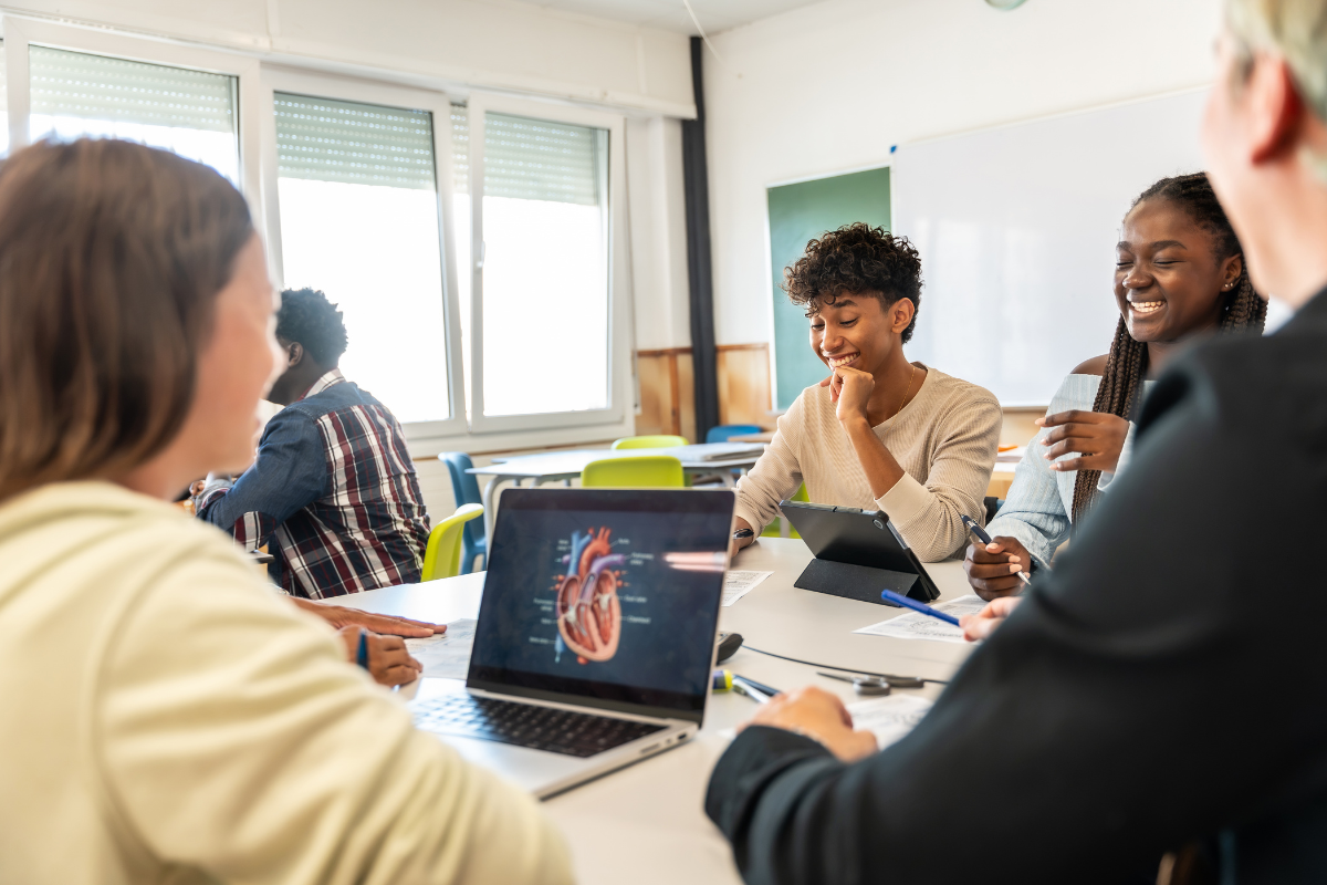 A bright classroom meeting where several students sit around a table using laptops and a tablet, smiling and talking about STEM teaching examples.