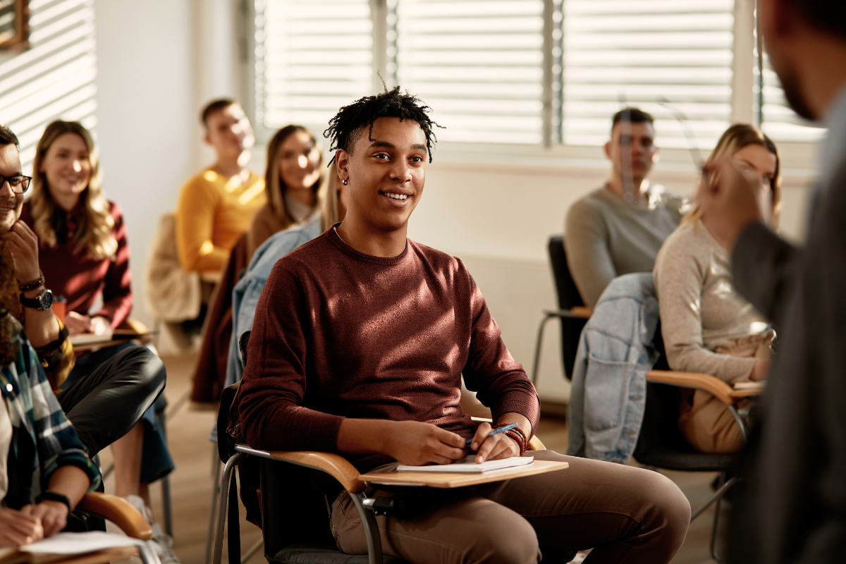 A young student with curly hair smiles while sitting in a classroom with other students attending a lecture designed using the student success rubric.
