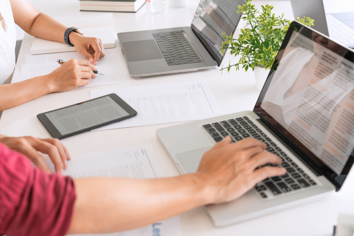 Faculty are working at a desk on accessible course materials with laptops, a tablet, and papers scattered around. One person is writing on a paper with a pen, while the other is typing on a laptop. A small potted plant is visible on the desk.