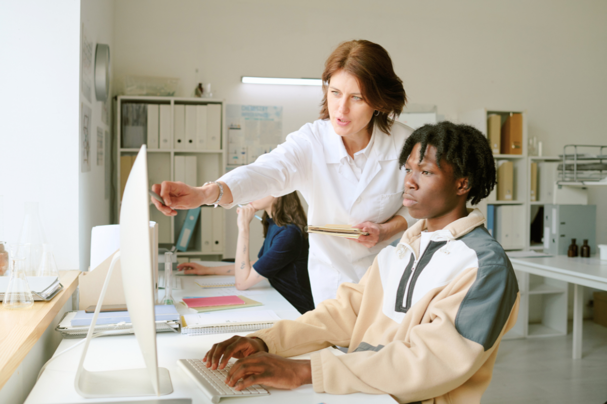 A female professor in a white lab coat guides a student in a beige sweater while they work at a computer in a bright gateway STEM course lab.