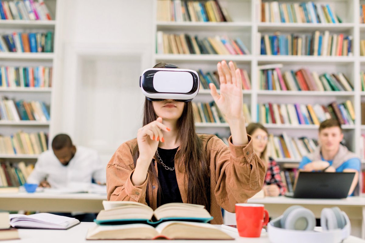 A student wearing a VR headset for immersive pedagogy raises their hand in a library setting with bookshelves in the background.